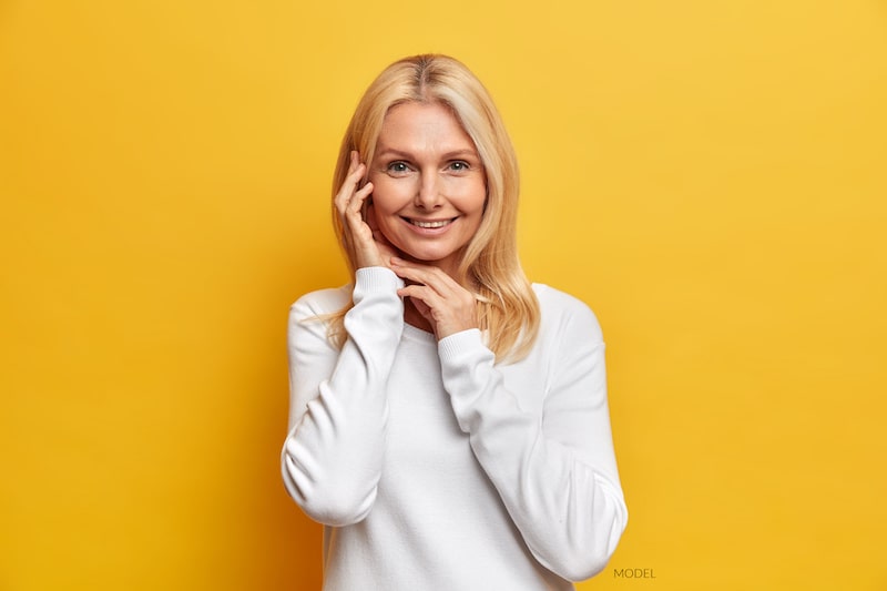 Smiling, middle-aged woman wearing a white shirt and standing with yellow background.