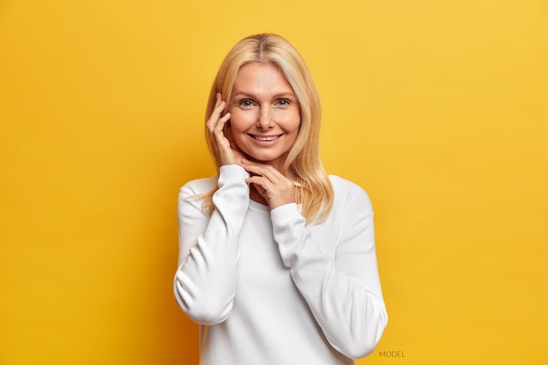 Smiling, middle-aged woman wearing a white shirt and standing  with yellow background.